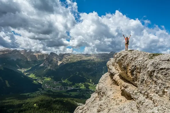 Person steht auf einem hohen Felsen mit ausgebreiteten Armen, überblickt eine weite Berglandschaft unter einem bewölkten Himmel